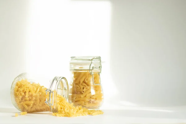 Pasta in a glass jar on a white background. Two containers stand on a table in daylight, a shadow falls from the dishes. One dishware is tipped over, contents spilled out of it. Safe food storage for future use.