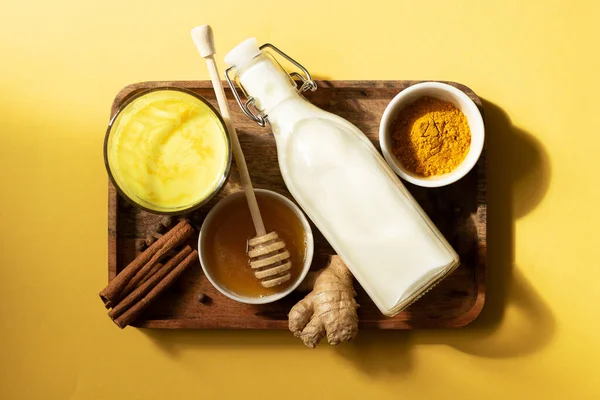 Kurkuma-Milch und die Zutaten für das Getränk liegen auf einem Holztablett. Gelber Hintergrund und Draufsicht. Natürliches Licht und Schatten — Stockfoto