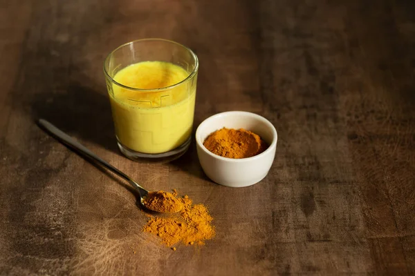 Goldene Milch in einer Glasschale. Daneben gibt es Kurkuma zur Zubereitung eines Getränks und einen Löffel mit Gewürzen. Dunkler Hintergrund — Stockfoto