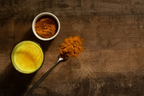 Goldene Milch in einer Glasschale. Daneben gibt es Kurkuma zur Zubereitung eines Getränks und einen Löffel mit Gewürzen. Dunkler Hintergrund — Stockfoto