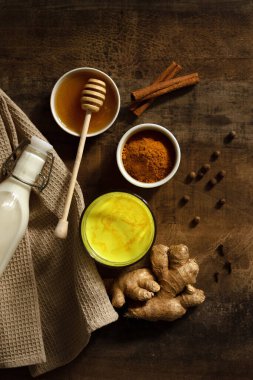 Zerdeçal sütü bardağa dökülür. Yanında içki yapmak için gereken malzemeler var. Peçetedeki süt, baharat ve bal. Karanlık huysuz arka planda altın içecek, seçici odaklanma. Üst görünüm.