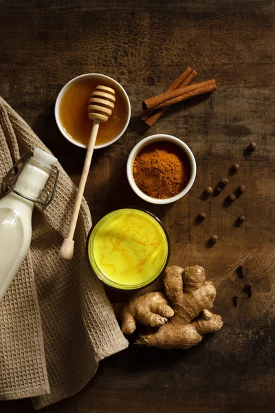 Zerdeçal sütü bardağa dökülür. Yanında içki yapmak için gereken malzemeler var. Peçetedeki süt, baharat ve bal. Karanlık huysuz arka planda altın içecek, seçici odaklanma. Üst görünüm.