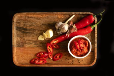 Sos kabına Harissa sosu. Pişirmek için gerekenler: Biber, sarımsak ve baharat. Her şey ahşap bir tepside yatıyor. Siyah arkaplan ve üst görünüm.