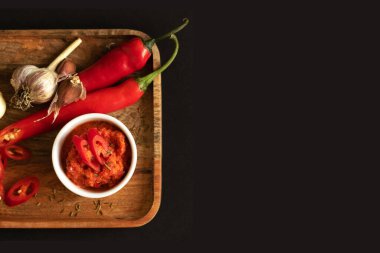 Sos kabına Harissa sosu. Pişirmek için gerekenler: Biber, sarımsak ve baharat. Her şey ahşap bir tepside yatıyor. Siyah arkaplan ve üst görünüm.