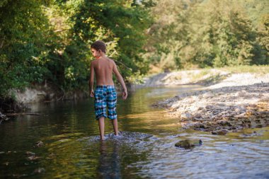 A teenage boy in shorts plays in a mountain river in shallow water in summer clipart