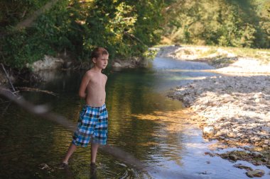 A teenage boy in shorts plays in a mountain river in shallow water in summer clipart