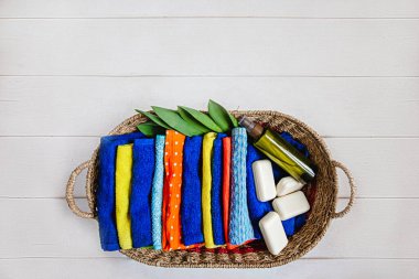 Bir sepet dolusu renkli Terry havlusu, birkaç parça beyaz sabun ve bir şişe duş jeli. Havluların yanında çiçeklerden taze yeşil yapraklar var. Tepeden manzara.