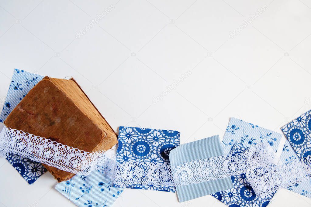 Square patches of blue printed fabric, old shebby book and white lace on a white background. Flat lay with space for text