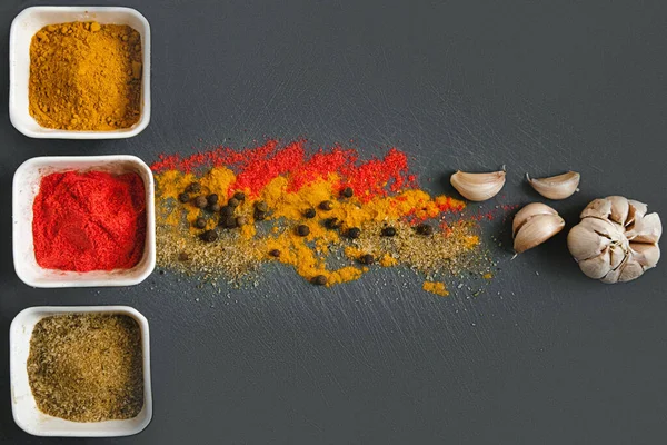 Three white square jars of various spices on a gray chopping Board. Next to them are scattered spices and garlic