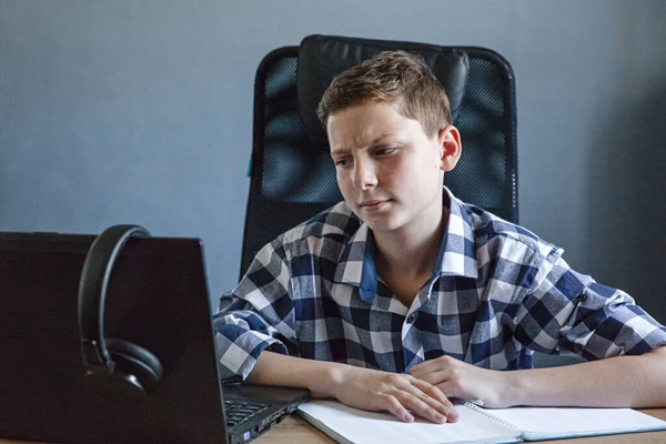 Evde dizüstü bilgisayarla tişört giyen genç bir çocuk ders çalışıyor. Masanın üzerinde açık bir defter ile oturuyor. Kulaklıklar laptopta asılı. Uzaklık öğrenme başlığı