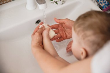 Anne, küçük oğlunun ellerini lavaboda yıkamasına yardım ediyor.