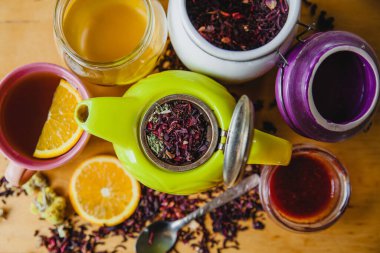 Yeşil çaydanlığı çay yaprakları ve otlarla birlikte aç. Etrafında bal, yaprak çayı ve reçel kavanozlarda. Yanında da bir fincan çay, portakal, kaşık ve dağınık çay yaprakları..