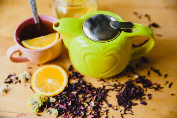 Grüne Teekanne auf einem Holztisch. Neben einer Scheibe Orange, einer vollen Tasse Tee, einem Honigglas und verstreuten Teeblättern und Kräutern. — Stockfoto