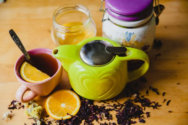 Green teapot on a wooden table. Next to a slice of orange, full cup of tea, honey jar and scattered tea leaves and herbs. — Stock Photo, Image