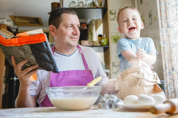 A father with a young son at the kitchen table at home preparing dough. Dad is reading a book, and the boy is busy in the flour.