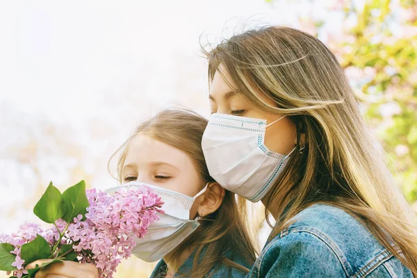 Mom and daughter in medical masks sniff a lilac on the street. Quarantine exit concept