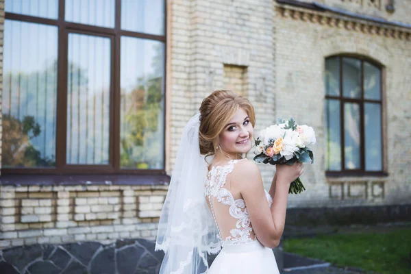 A noiva em um belo vestido de noiva com um laço aberto de volta com um véu longo. A noiva está segurando um buquê de casamento. Noiva com um belo sorriso — Fotografia de Stock