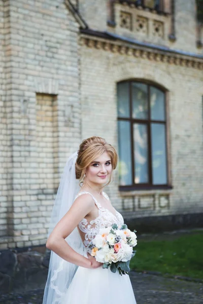 A noiva em um belo vestido de noiva com um laço aberto de volta com um véu longo. A noiva está segurando um buquê de casamento. Noiva com um belo sorriso — Fotografia de Stock