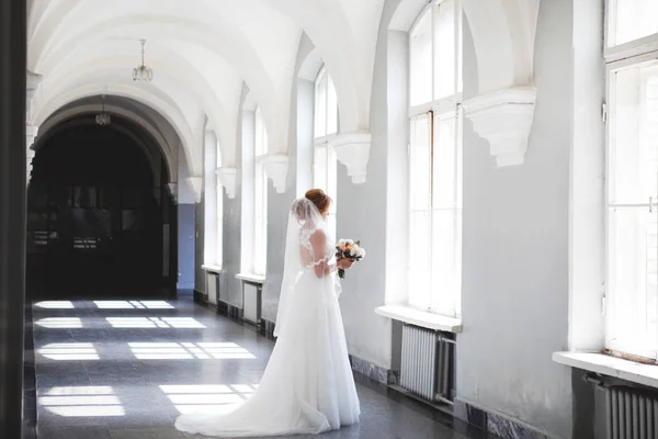 A noiva em um lindo vestido de noiva com um longo véu fica na frente de uma grande janela e olha para fora da janela. Noiva em um quarto com arcos brancos altos — Fotografia de Stock