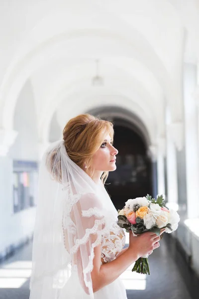 Невеста в красивом свадебном платье с длинной вуалью стоит перед большим окном и смотрит в окно. Невеста в комнате с высокими белыми арками — стоковое фото