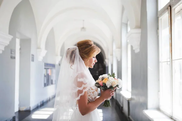 Невеста в красивом свадебном платье с длинной вуалью стоит перед большим окном и смотрит на красивый свадебный букет. Невеста в помещении с белыми арками смотрит на свадебный букет в ней — стоковое фото