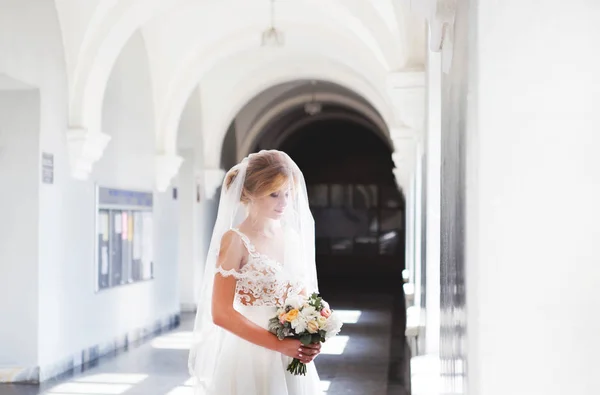 A noiva em um lindo vestido de noiva com um longo véu fica na frente de uma grande janela e olha para baixo. O olhar da noiva para baixo. Pintar a sombra do sol cai no chão das janelas . — Fotografia de Stock