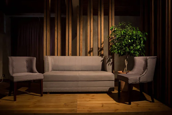 Кинематографический свет. Серый диван и два серых стула на деревянных ножках стоят на коричневом деревянном подиуме, за вертикальными деревянными балками и большим деревянным горшком — стоковое фото