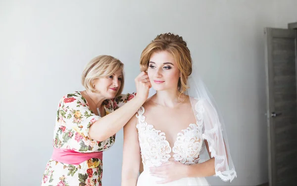 Manhã da noiva. Mamãe ajuda a colocar os brincos de uma noiva bonita. Mamãe corrige o penteado do casamento da noiva. Noiva em um lindo vestido de noiva — Fotografia de Stock