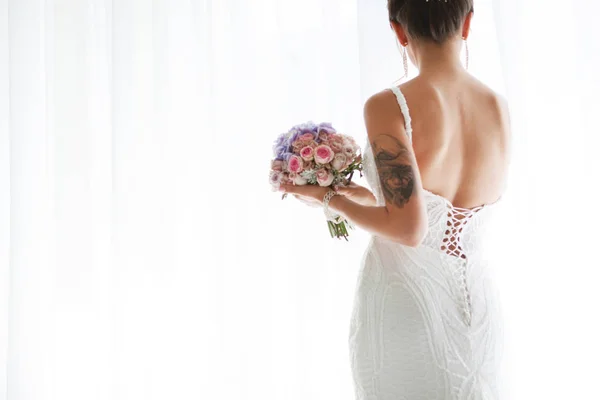 Steht die Braut mit dem Rücken zum Fenster in einem wunderschönen Brautkleid. die Braut hält einen schönen Strauß in der Hand. Braut mit Tätowierung auf der Hand — Stockfoto