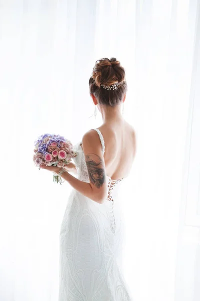 La mariée se tient avec son dos à la fenêtre dans une belle robe de mariée. La mariée tient un beau bouquet. Mariée avec tatouage à portée de main — Photo