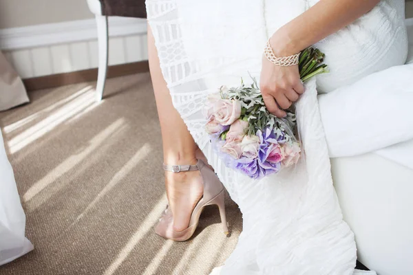 Die Braut, die am Hochzeitsmorgen auf einem Bett sitzt. Hochzeitskleid und nudefarbene Schuhe. mit einem Brautstrauß in der Hand — Stockfoto