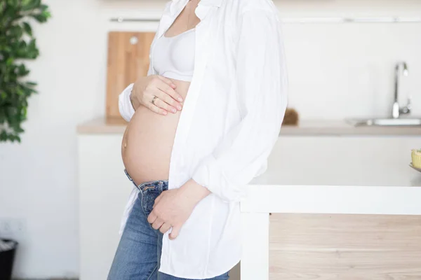 Une femme enceinte se tient dans la cuisine en jeans et une chemise blanche — Photo