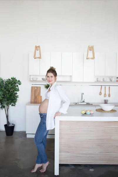 Kobiety w ciąży stoi w kuchni w dżinsy i koszulę — Zdjęcie stockowe