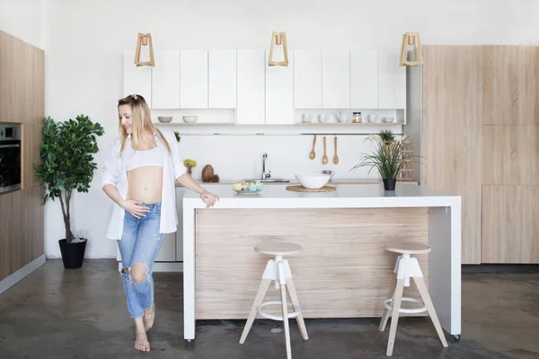 Беременная женщина стоит на кухне в джинсах и белой рубашке — стоковое фото
