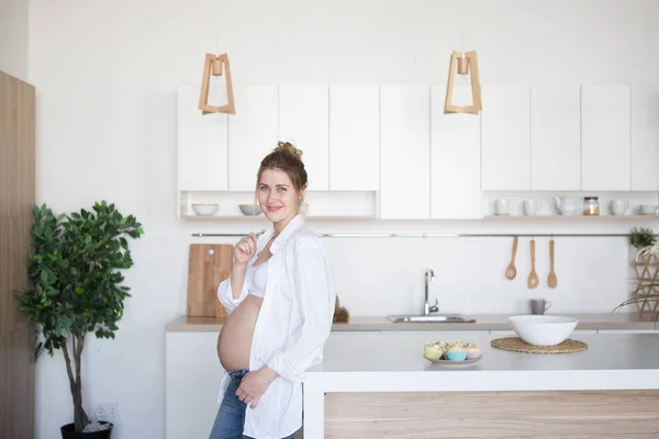 妊娠中の女性はジーンズと白いシャツで台所に立っています。 — ストック写真
