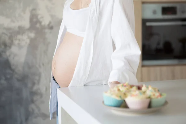 Une femme enceinte se tient dans la cuisine en jeans et une chemise blanche. Dans le cadre, une assiette avec des gâteaux — Photo
