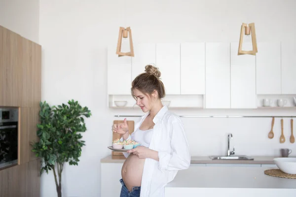 임신한 여자는 부엌에서 케이크 접시 보유 하고있다. 임산부는 청바지와 흰 셔츠에 부엌에 서 서 — 스톡 사진