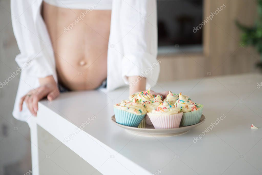 A pregnant woman is standing in the kitchen in jeans and a white shirt. In the frame, a plate with cakes