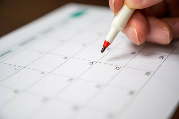 The hand of a man holding a pen in his hand and recording his schedule on a desk calendar.