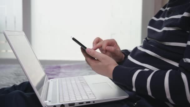 Un hombre asiático de mediana edad teletransportándose desde casa en un portátil está comprobando algo con su teléfono inteligente en la mano . — Vídeo de stock