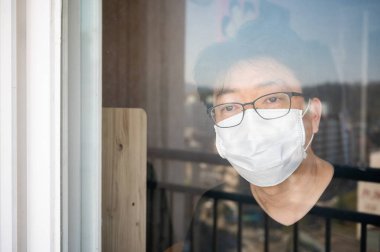 COVID-19 coronavirus self-isolation concept. An Asian man with a face mask and a window looking out the window during his self-isolation at home. clipart