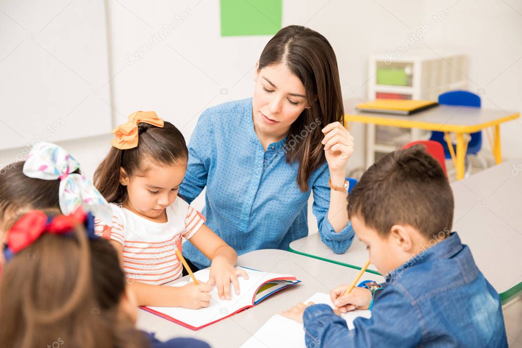 Pretty young preschool teacher helping her pupils with some writing assigment in the classroom