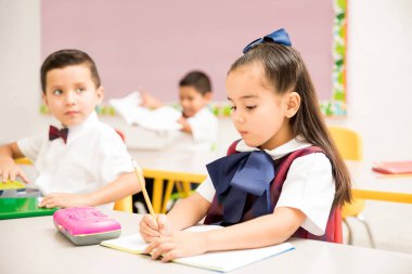 Cute preschool students wearing a uniform and doing a writing assigment in a classroom clipart