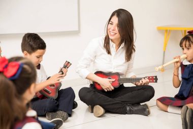 Fun female preschool teacher playing a guitar and teaching some music to her students clipart