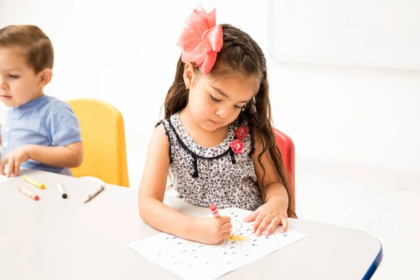 Beautiful little girl in a kindergarten class using crayons to color a drawing in the classroom