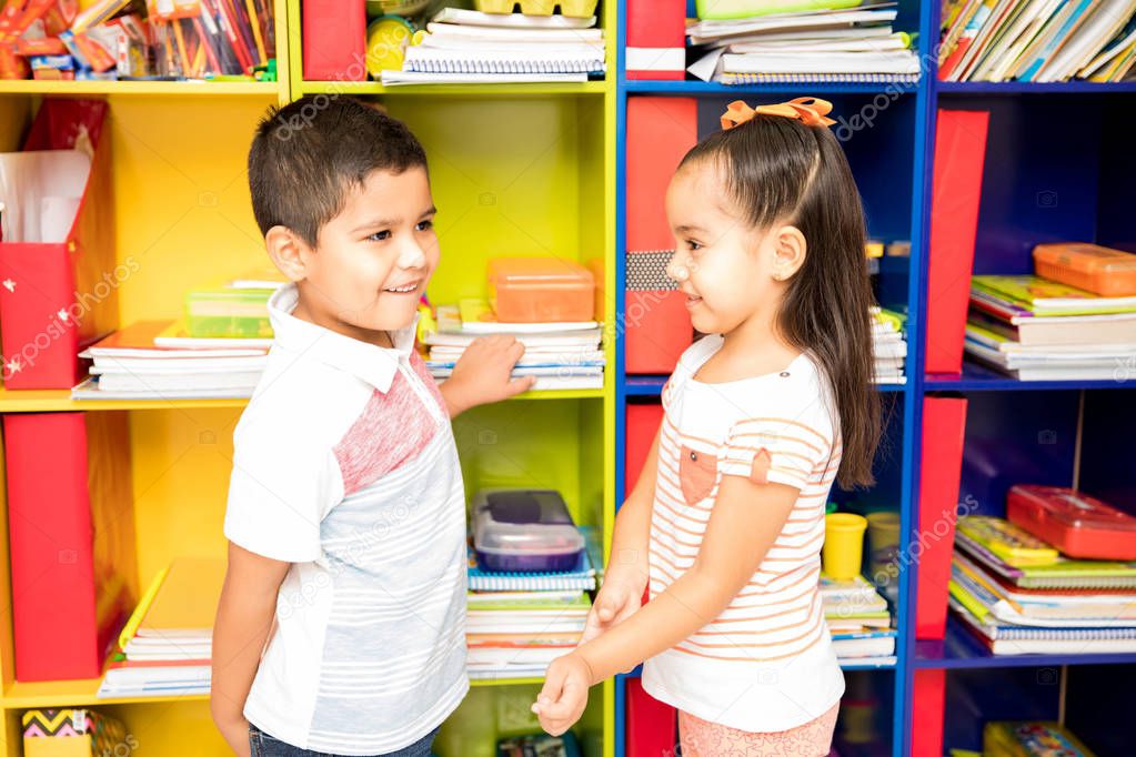 Profile view of a shy little boy and a girl flirting with each other while standing in front of a classroom locker