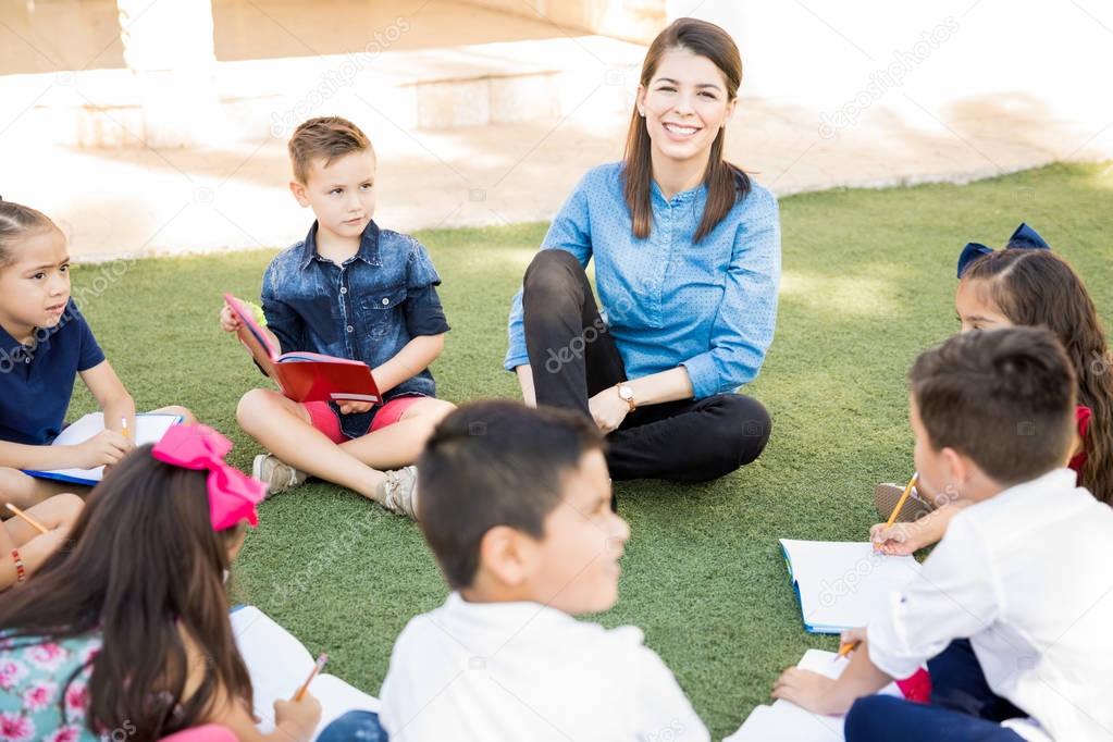 Pretty Hispanic preschool teacher taking her students outside and having a fun class on the grass