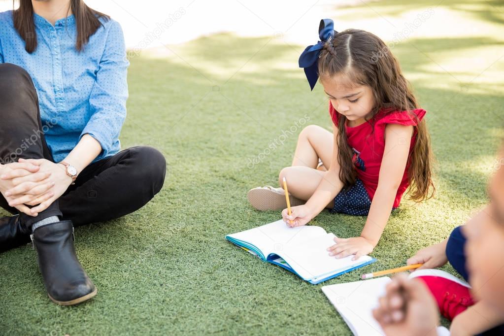 Cute little girl doing a writing assigment while sitting on the grass outdoors 