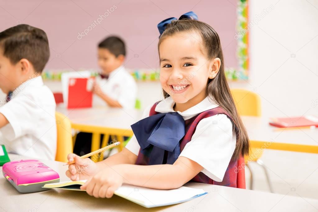 Portrait of a gorgeous Latin preschool student in uniform doing a writing assigment in a classroom and smiling