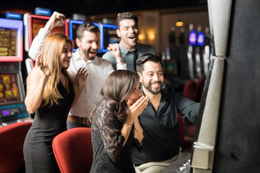 Beautiful Hispanic woman looking excited about hitting the jackpot in a slot machine while her friends celebrate with her clipart